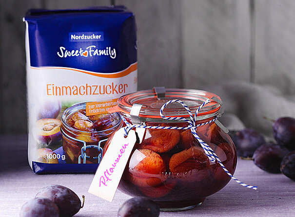 Eine Packung Einmachzucker von Sweetfamily, davor ein bauchiges Glas mit eingemachten Pflaumen.